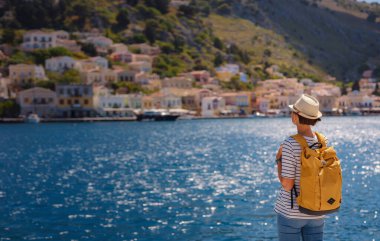 Sırt çantalı mutlu Asyalı kadın Symi Adaları 'nda tatilinin tadını çıkarıyor. Liman manzarası, Yunanistan 'ın Dodecanese kentindeki küçük bir ada, sakin bir atmosfer ve muhteşem bir mimari..