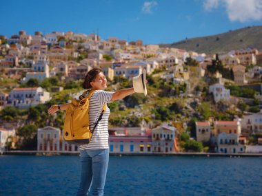 Sırt çantalı mutlu Asyalı kadın Symi Adaları 'nda tatilinin tadını çıkarıyor. Liman manzarası, Yunanistan 'ın Dodecanese kentindeki küçük bir ada, sakin bir atmosfer ve muhteşem bir mimari..