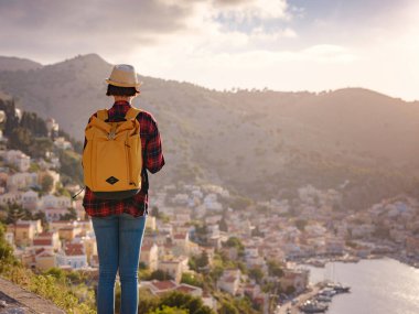 Sırt çantalı mutlu Asyalı kadın gün batımında Symi Adaları 'nda tatilin tadını çıkarıyor. Liman manzarası, Yunanistan 'ın Dodecanese kentindeki küçük bir ada, sakin bir atmosfer ve muhteşem bir mimari..