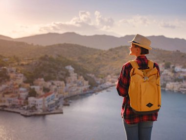 Sırt çantalı mutlu Asyalı kadın gün batımında Symi Adaları 'nda tatilin tadını çıkarıyor. Liman manzarası, Yunanistan 'ın Dodecanese kentindeki küçük bir ada, sakin bir atmosfer ve muhteşem bir mimari..