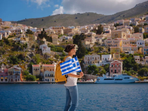 Sırt çantalı mutlu Asyalı kadın ve Yunan bayrağı Symi Adaları 'nda tatilinin tadını çıkarıyor. Liman manzarası, Yunanistan 'ın Dodecanese kentindeki küçük bir ada, sakin bir atmosfer ve muhteşem bir mimari..
