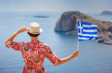 Yunan Adaları 'nda güneşli bir günün tadını çıkaran güzel mutlu kadın. Yunanistan 'a seyahat, turizm sezonu dışında Akdeniz adaları. Ulusal Yunan bayrağıyla gezen genç kadın deniz manzarasının keyfini çıkarıyor