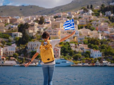 Sırt çantalı mutlu Asyalı kadın ve Yunan bayrağı Symi Adaları 'nda tatilinin tadını çıkarıyor. Liman manzarası, Yunanistan 'ın Dodecanese kentindeki küçük bir ada, sakin bir atmosfer ve muhteşem bir mimari..