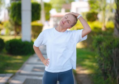 Modern bir şehirde sağlıklı yaşam tarzı ve spor fikri. Kadın koşucu. Genç beyaz kadın koşmadan önce parkta kaslarını esnetiyor ve dışarıda sağlıklı bir şekilde eğleniyor. Kamu parkındaki fitness kızı.