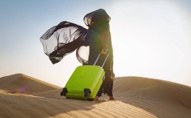 Geleneksel abaya kostümlü, açık yeşil ya da sarı kumlu bavulu olan Arap bir kadın. Birleşik Arap Emirlikleri 'ne seyahat kavramı ve fikri, gün batımında Dubai çölündeki kum tepeleri.