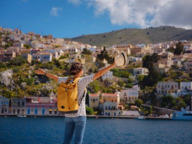 Sırt çantalı mutlu Asyalı kadın Symi Adaları 'nda tatilinin tadını çıkarıyor. Liman manzarası, Yunanistan 'ın Dodecanese kentindeki küçük bir ada, sakin bir atmosfer ve muhteşem bir mimari..