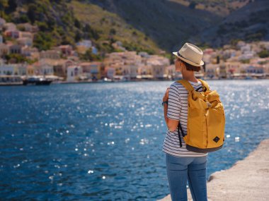 Sırt çantalı mutlu Asyalı kadın Symi Adaları 'nda tatilinin tadını çıkarıyor. Liman manzarası, Yunanistan 'ın Dodecanese kentindeki küçük bir ada, sakin bir atmosfer ve muhteşem bir mimari..
