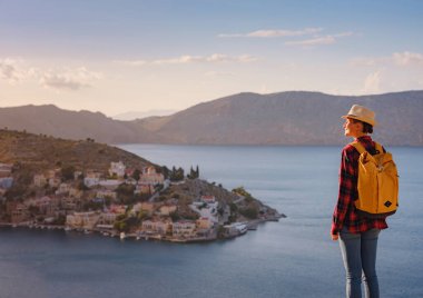 Sırt çantalı mutlu Asyalı kadın gün batımında Symi Adaları 'nda tatilin tadını çıkarıyor. Liman manzarası, Yunanistan 'ın Dodecanese kentindeki küçük bir ada, sakin bir atmosfer ve muhteşem bir mimari..