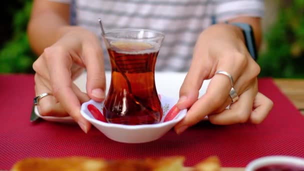 女性は庭で伝統的なトルコ式朝食を楽しんでいます 彼女は緑に囲まれたテーブルに座っている トルコの伝統的なティーカップで紅茶に砂糖をかき混ぜる女性 — ストック動画