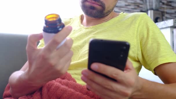 薬を服用するためのスマートフォン研究の指示を使用して男 病気の男はリビングでソファーに座っている ホーム医学コンセプト インフルエンザ救助錠剤 医療アドバイス — ストック動画