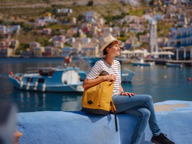 Sırt çantalı mutlu Asyalı kadın Symi Adaları 'nda tatilinin tadını çıkarıyor. Liman manzarası, Yunanistan 'ın Dodecanese kentindeki küçük bir ada, sakin bir atmosfer ve muhteşem bir mimari..