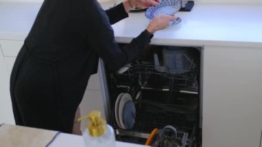 Beyaz, modern mutfakta ev ödevi yapan bir kadın. Kadın bulaşık makinesine kirli bulaşık koyuyor.