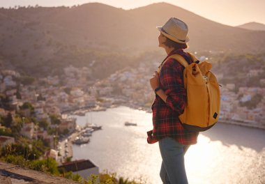 Sırt çantalı mutlu Asyalı kadın gün batımında Symi Adaları 'nda tatilin tadını çıkarıyor. Liman manzarası, Yunanistan 'ın Dodecanese kentindeki küçük bir ada, sakin bir atmosfer ve muhteşem bir mimari..