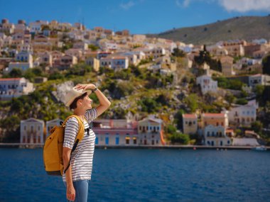 Sırt çantalı mutlu Asyalı kadın Symi Adaları 'nda tatilinin tadını çıkarıyor. Liman manzarası, Yunanistan 'ın Dodecanese kentindeki küçük bir ada, sakin bir atmosfer ve muhteşem bir mimari..