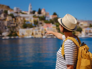 Sırt çantalı mutlu Asyalı kadın Symi Adaları 'nda tatilinin tadını çıkarıyor. Liman manzarası, Yunanistan 'ın Dodecanese kentindeki küçük bir ada, sakin bir atmosfer ve muhteşem bir mimari..