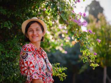 Genç Asyalı kadın güneşli yeşil parkta duruyor, çiçek açan bougainvillea çalısına hayran. Çiçeklerin parlak renkleri parkların yemyeşil renklerinin tam zıttıdır..