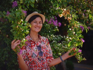 Genç Asyalı kadın güneşli yeşil parkta duruyor, çiçek açan bougainvillea çalısına hayran. Çiçeğin parlak renkleri, parkın yemyeşil renklerinin çok güzel bir tezatıdır..