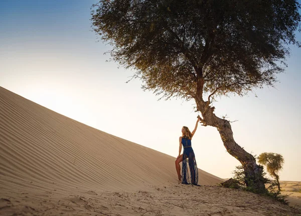 Desert adventure. Young arabian Woman in sexy boho blue makrame dress in sands dunes of UAE desert at sunset. The Dubai Desert Conservation Reserve, United Arab Emirates.