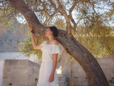 Açık havada, zeytin ağacının yanında beyaz elbiseli güzel bir Asyalı genç kadın. Temiz havayı kucaklamak ve açık hava aktivitelerine katılmak. Friluftsliv konsepti, dışarıda mümkün olduğunca çok zaman geçirmek demektir.