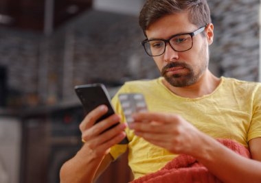 Genç beyaz adam okumak için akıllı telefon kullanıyor, ilaç reçetesini arıyor, internetten bilgi hakkında hap etiketi metni yazıyor, talimatlar yan etkiler, eczane tıbbı konsepti