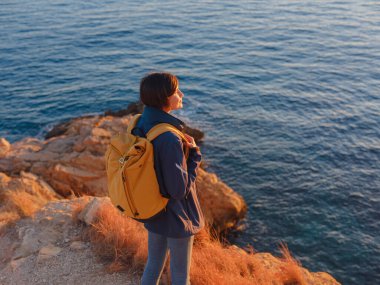 İspanya, Benidorm 'da kayalık sahilde yürüyüş yapan genç bir kadın. Dalgalı denizi ve körfezi izliyorum. Gezgin, huzurlu doğa manzarasında özgürlüğün tadını çıkarıyor.