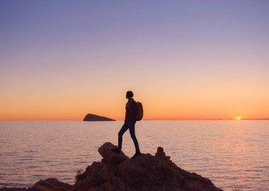 İspanya, Benidorm 'da kayalık plajda yürüyüş yapan genç bir kadının silueti. Dalgalı denizi ve körfezi izliyorum. Huzurlu günbatımı manzarasında özgürlüğün tadını çıkaran gezgin, başarı konsepti