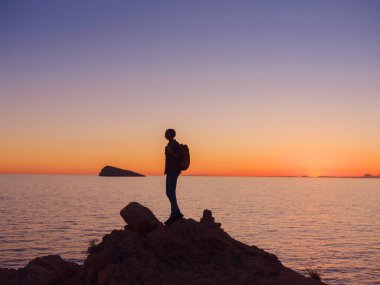 İspanya, Benidorm 'da kayalık plajda yürüyüş yapan genç bir kadının silueti. Dalgalı denizi ve körfezi izliyorum. Huzurlu günbatımı manzarasında özgürlüğün tadını çıkaran gezgin, başarı konsepti