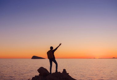 İspanya, Benidorm 'da kayalık plajda yürüyüş yapan genç bir kadının silueti. Dalgalı denizi ve körfezi izliyorum. Huzurlu günbatımı manzarasında özgürlüğün tadını çıkaran gezgin, başarı konsepti