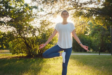 Genç Asyalı bir kadın sabah ya da akşam parkta yoga yapıyor sağlıklı bir kadın şehir parkında ağaç pozu veriyor. Farkındalık, kararlılık, sağlıklı alışkanlıklar ve denge konsepti