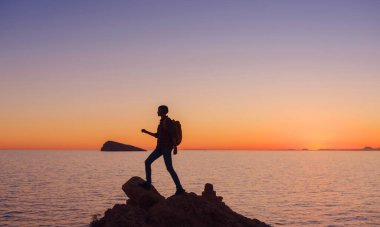 İspanya, Benidorm 'da kayalık plajda yürüyüş yapan genç bir kadının silueti. Dalgalı denizi ve körfezi izliyorum. Huzurlu günbatımı manzarasında özgürlüğün tadını çıkaran gezgin, başarı konsepti