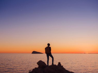 İspanya, Benidorm 'da kayalık plajda yürüyüş yapan genç bir kadının silueti. Dalgalı denizi ve körfezi izliyorum. Huzurlu günbatımı manzarasında özgürlüğün tadını çıkaran gezgin, başarı konsepti