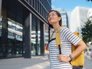 Viyana 'daki modern şehir mimarisinin arka planında sarı sırt çantalı Asyalı kadın. Düşünceli kadın hippi modern Viyana şehrinin manzarasına hayran.