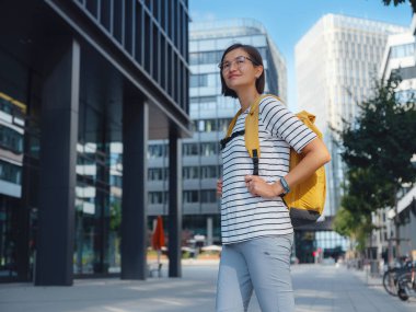 Viyana 'daki modern şehir mimarisinin arka planında sarı sırt çantalı Asyalı kadın. Düşünceli kadın hippi modern Viyana şehrinin manzarasına hayran.