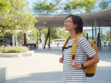 Viyana 'daki modern şehir mimarisinin arka planında sarı sırt çantalı Asyalı kadın. Düşünceli kadın hippi modern Viyana şehrinin manzarasına hayran.
