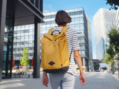 Viyana 'daki modern şehir mimarisinin arka planında sarı sırt çantalı Asyalı kadın. Düşünceli kadın hippi modern Viyana şehrinin manzarasına hayran.