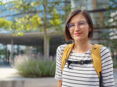 Viyana 'daki modern şehir mimarisinin arka planında sarı sırt çantalı Asyalı kadın. Düşünceli kadın hippi modern Viyana şehrinin manzarasına hayran.