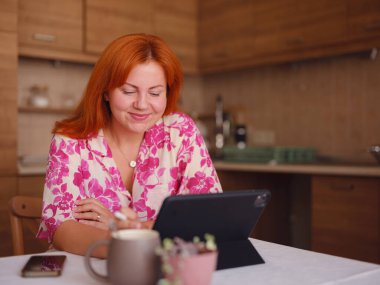 Genç bir kadın ev mutfağında tablet üzerinde çalışıyor. Gerçek bir serbest çalışan kadın bilgisayarını evden bir fincan kahveyle uzaktan çalışmak için kullanır..