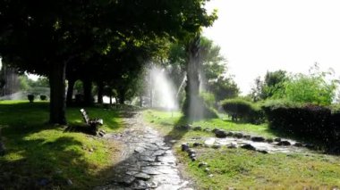 Antalyas yeşil parkında ferahlatıcı sahneler: fıskiyeler yemyeşil çimlere serpiştirir, şehrin kalbinde huzurlu bir vaha sunar. Çimenler yemyeşil, fıskiyeler havada sis yaratır..