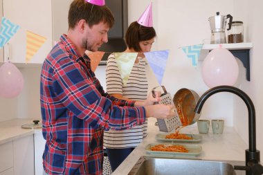 Genç çift sıcak bir ortamda pasta ve kahkahaların tadını çıkararak evde rahat bir doğum günü kutluyor. Neşeli atmosfer, renkli dekorasyonlar, şenlikli bir akşam yemeği pişirme, lezzetli makarna.