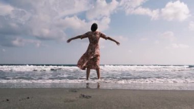 Uçan elbiseli bir kadın Akdeniz sahilinde Antalya, Türkiye 'de Antalya plajında yürürken, sıcak güneş ve hafif esintinin tadını çıkarıyor. Huzur ve memnuniyet içinde, sadece anın tadını çıkarıyor..