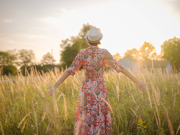Молодая женщина, идущая по живописному европейскому полю в конце лета. Золотой солнечный свет, пышная зелень и спокойная сельская атмосфера создают мирную сельскую местность.
