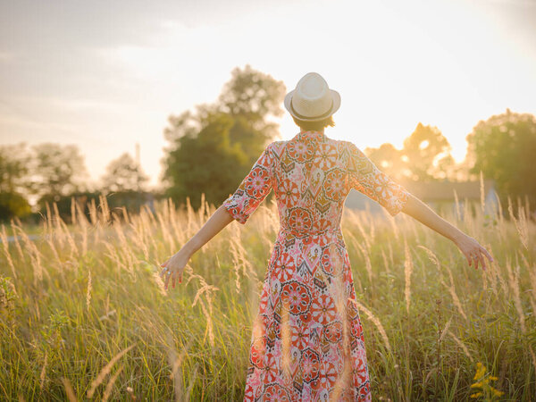 Молодая женщина, идущая по живописному европейскому полю в конце лета. Золотой солнечный свет, пышная зелень и спокойная сельская атмосфера создают мирную сельскую местность.