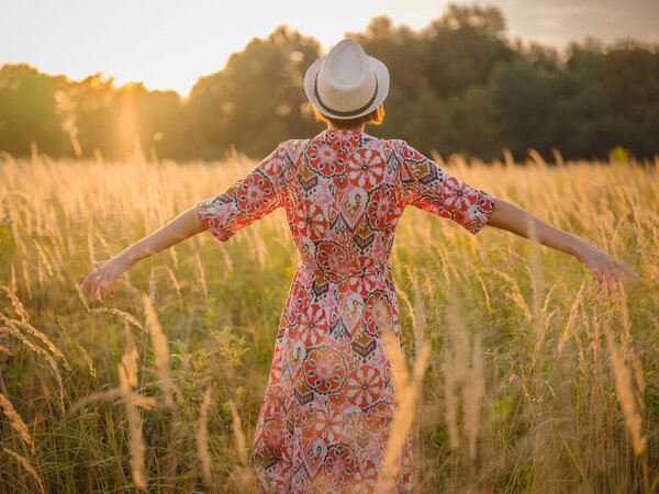 Молодая женщина, идущая по живописному европейскому полю в конце лета. Золотой солнечный свет, пышная зелень и спокойная сельская атмосфера создают мирную сельскую местность.