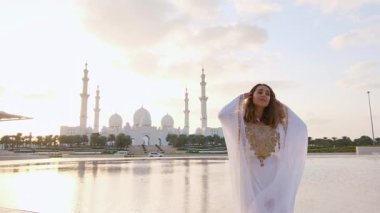 Abu Dabi 'deki Şeyh Zayed Büyük Camii yakınlarındaki Wahat Al Karama' daki geleneksel Arap elbiseli kadın, gün batımında kültürel zarafet ve ikonik İslami mimari sergiliyor..
