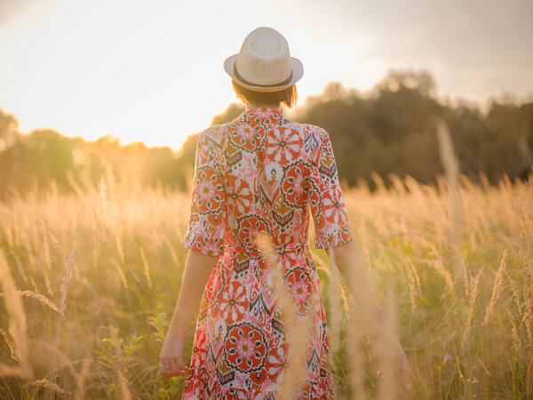 Молодая женщина, идущая по живописному европейскому полю в конце лета. Золотой солнечный свет, пышная зелень и спокойная сельская атмосфера создают мирную сельскую местность.
