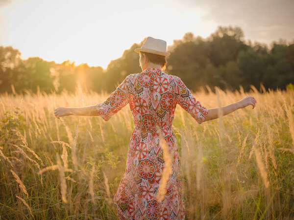 Молодая женщина, идущая по живописному европейскому полю в конце лета. Золотой солнечный свет, пышная зелень и спокойная сельская атмосфера создают мирную сельскую местность.