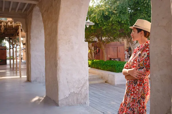 Dubai 'de Al Seef Parkı' nı gezen etnik kıyafetli ve şapkalı genç bir kadın. Geleneksel Emirati mimarisi ve modern atmosferiyle çevrelenmiş Dubai Deresi 'nin kültürel mirasının cazibesine sahip..