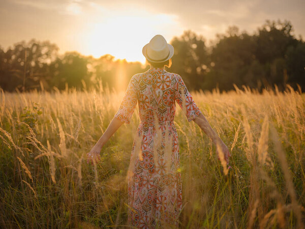 Молодая женщина, идущая по живописному европейскому полю в конце лета. Золотой солнечный свет, пышная зелень и спокойная сельская атмосфера создают мирную сельскую местность.