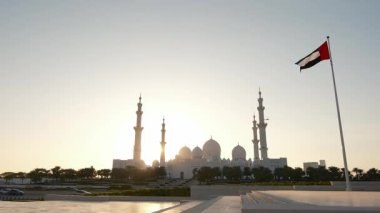 Wahat Al Karama Parkı 'nda gün batımı, BAE bayrağı gururla dalgalanır, altın ışıkla aydınlanır. Arka planda sakin bir manzara ve Şeyh Zayed Camii çarpıcı ve vatansever bir manzara yaratır..