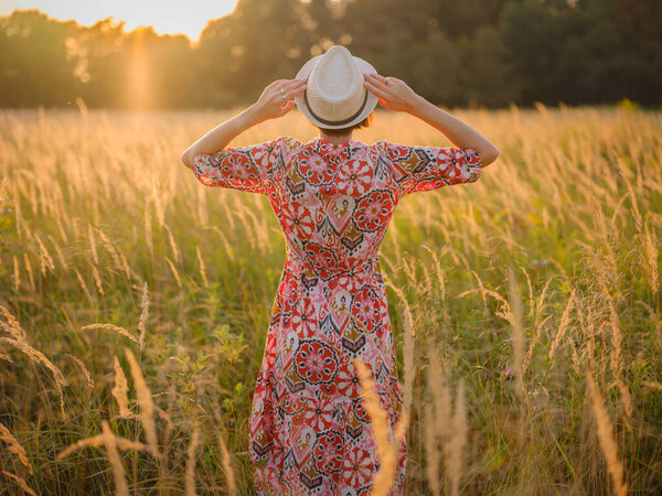 Молодая женщина, идущая по живописному европейскому полю в конце лета. Золотой солнечный свет, пышная зелень и спокойная сельская атмосфера создают мирную сельскую местность.